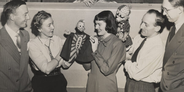 Five smiling people stand in a row, the third and fourth holding puppets