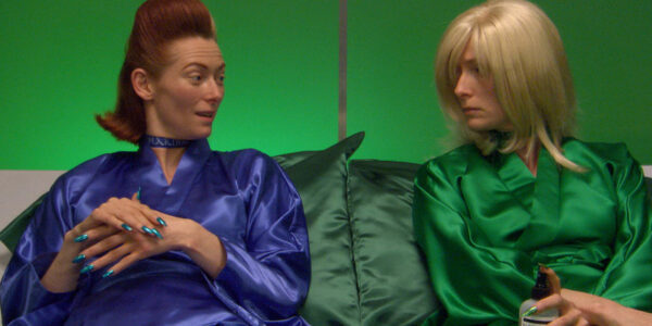 Two women—different versions of Tilda Swinton—sit on a couch and look at one another, one in purple and the other in green.