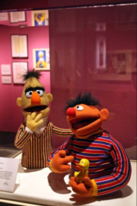 The puppets for the Sesame Street characters Bert and Ernie on display in a glass case at MoMI. Ernie is holding a rubber duckie.
