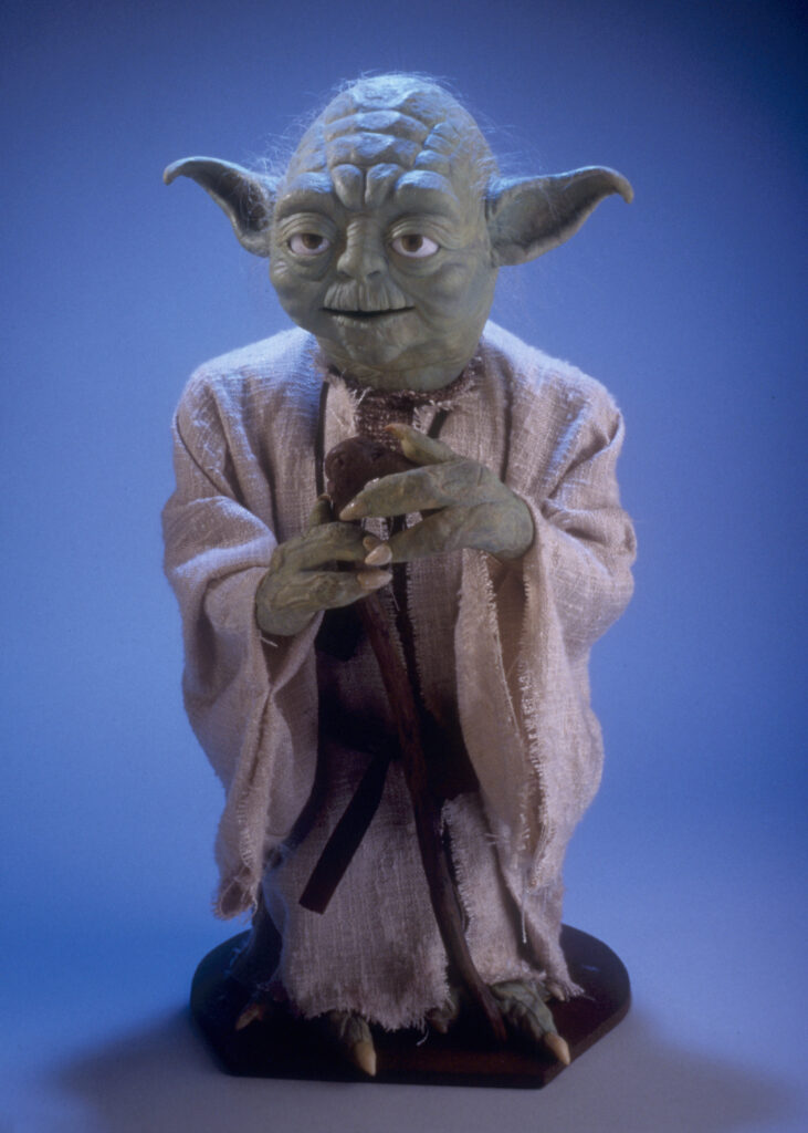A full portrait of a puppet of Yoda from Star Wars, holding a cane