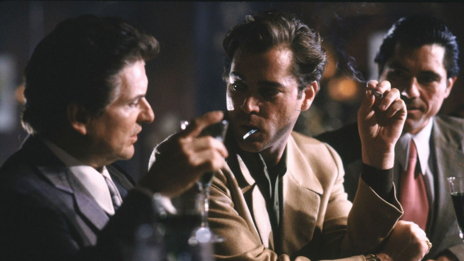 Two tough-looking gangsters sit at a bar, the one on right with a cigarette in his mouth