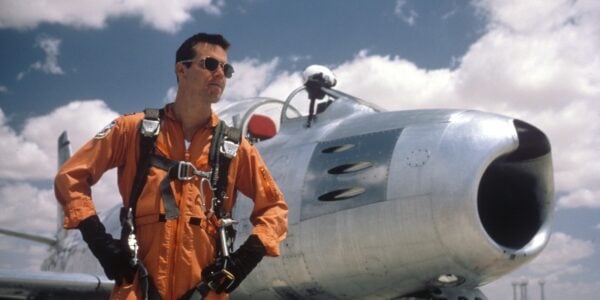 A test pilot in orange and aviator sunglasses standing in front of an airplane
