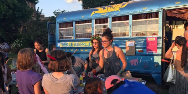 Two young women interacting with kids outside of a big blue bus.