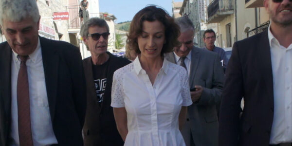 A woman in a white dress walking down a city street, flanked by men in suits