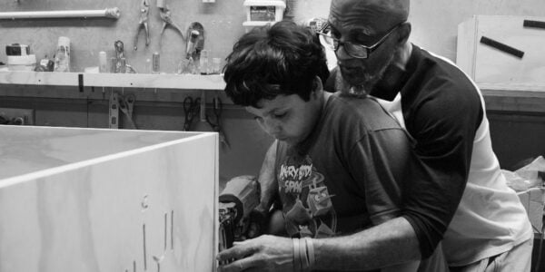 An older man has his arms around a young boy as he teaches him carpentry