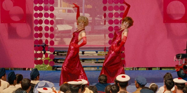 Two women in bright red dresses and silk gloves perform in sync on a stage with one arm raised to sky
