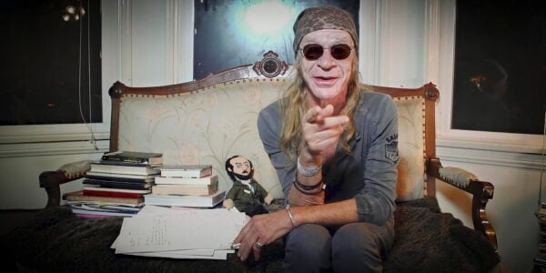 A man with long hair and in sunglasses sits on a couch and points at the camera