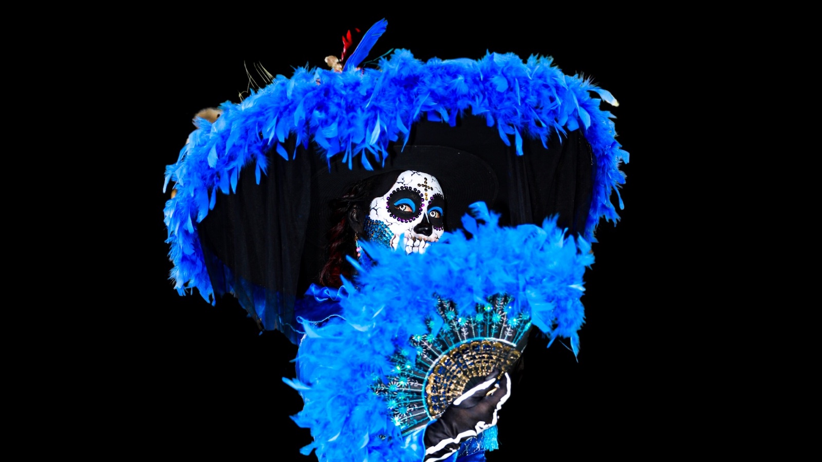 A person in a skull mask and feathery blue costume and holding a hand-fan, celebrates the Day of the Dead.