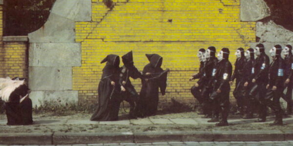 A group of hooded monks pull a rope in front of a yellow brick wall.