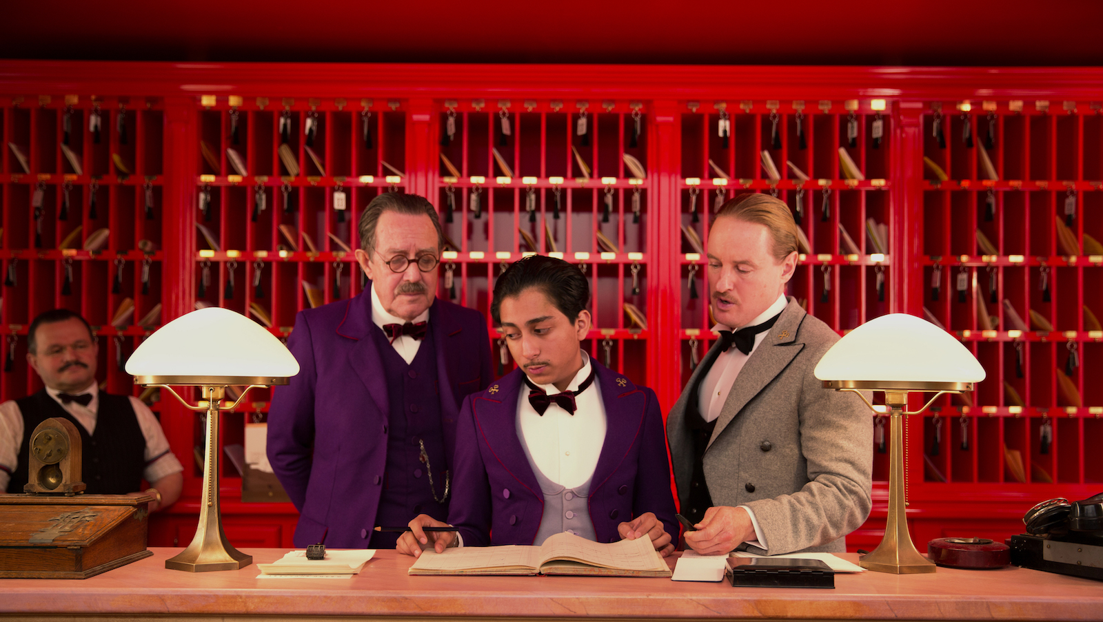 A young hotel bellboy flanked by two older mustachioed men against a red front desk backdrop