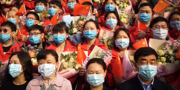A crowd of face-masked people in China look towards the camera, many holding flowers and wearing red.