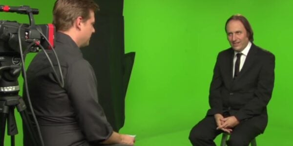 Two men sit on stools against a green-screen background
