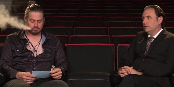 Two men in the front row of a theater, the one on the left exhaling smoke and reading a card in his hand.