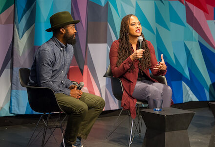 A man in a hat and a woman in a maroon jacket—cinematographer Bradford Young and director Ava DuVernay—speak to each other on-stage at Museum of the Moving Image, before a brightly colored kaleidoscopic curtain.