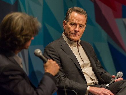 The actor Bryan Cranston intensely looks at his conversation partner on the Museum theater stage, in front of a brightly colored curtain.