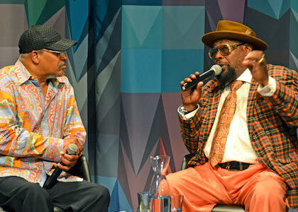 Two men in hats, musician George Clinton and journalist James Mtume, talk together into microphones in front of the multicolored curtain on the Museum stage.