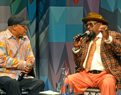 Two men in hats, musician George Clinton and journalist James Mtume, talk together into microphones in front of the multicolored curtain on the Museum stage.