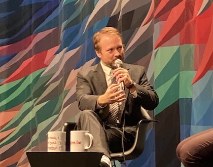 A man in a suit, director Rian Johnson, speaks into a mic on the Museum stage in front of the multicolored curtain.