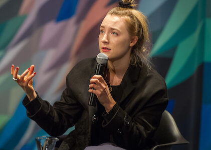 A young woman, actress Saoirse Ronan, gesticulates with her hands while talking on the Museum stage in front of the multicolored curtain.