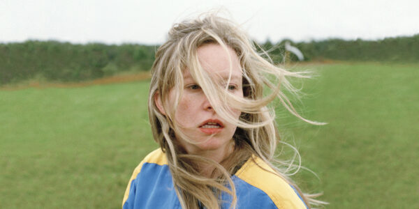 A distressed young woman stands in a field with long windswept blonde hair in her face.