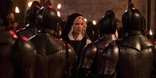 A nun in her habit is surrounded by guardsmen in dark armor.