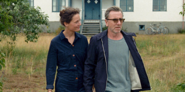 A woman in blue and a man in sunglasses walk outside away from a house toward the camera.
