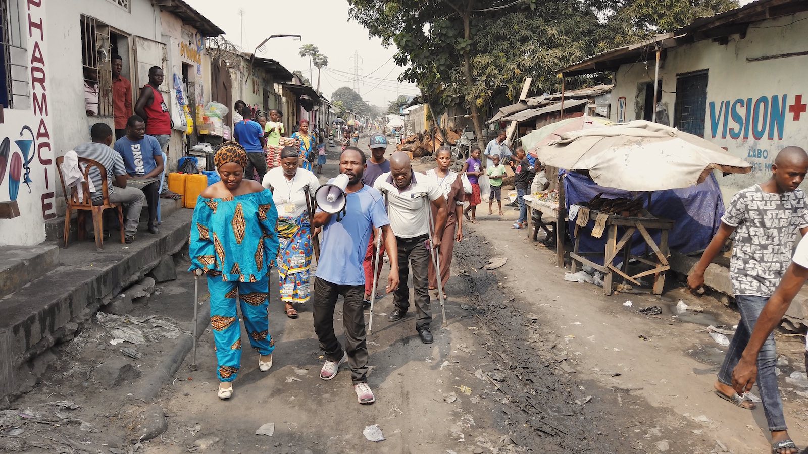 A group of people, one holding a megaphone and others in crutches, walk down a village street in Kinshasa.