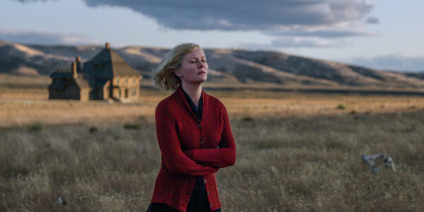 A woman (Kirsten Dunst) in a red sweater stands with her arms crossed against a vast Montana mountain landscape.