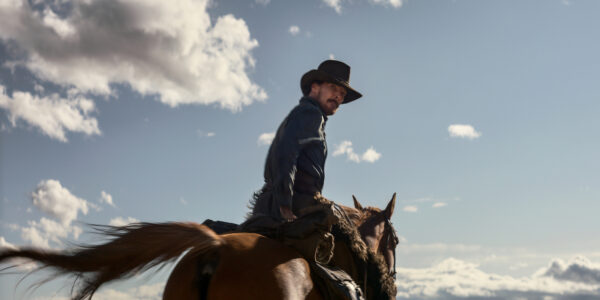 A man on horseback in a cowboy hat looks back over his shoulder, a blue horizon behind him