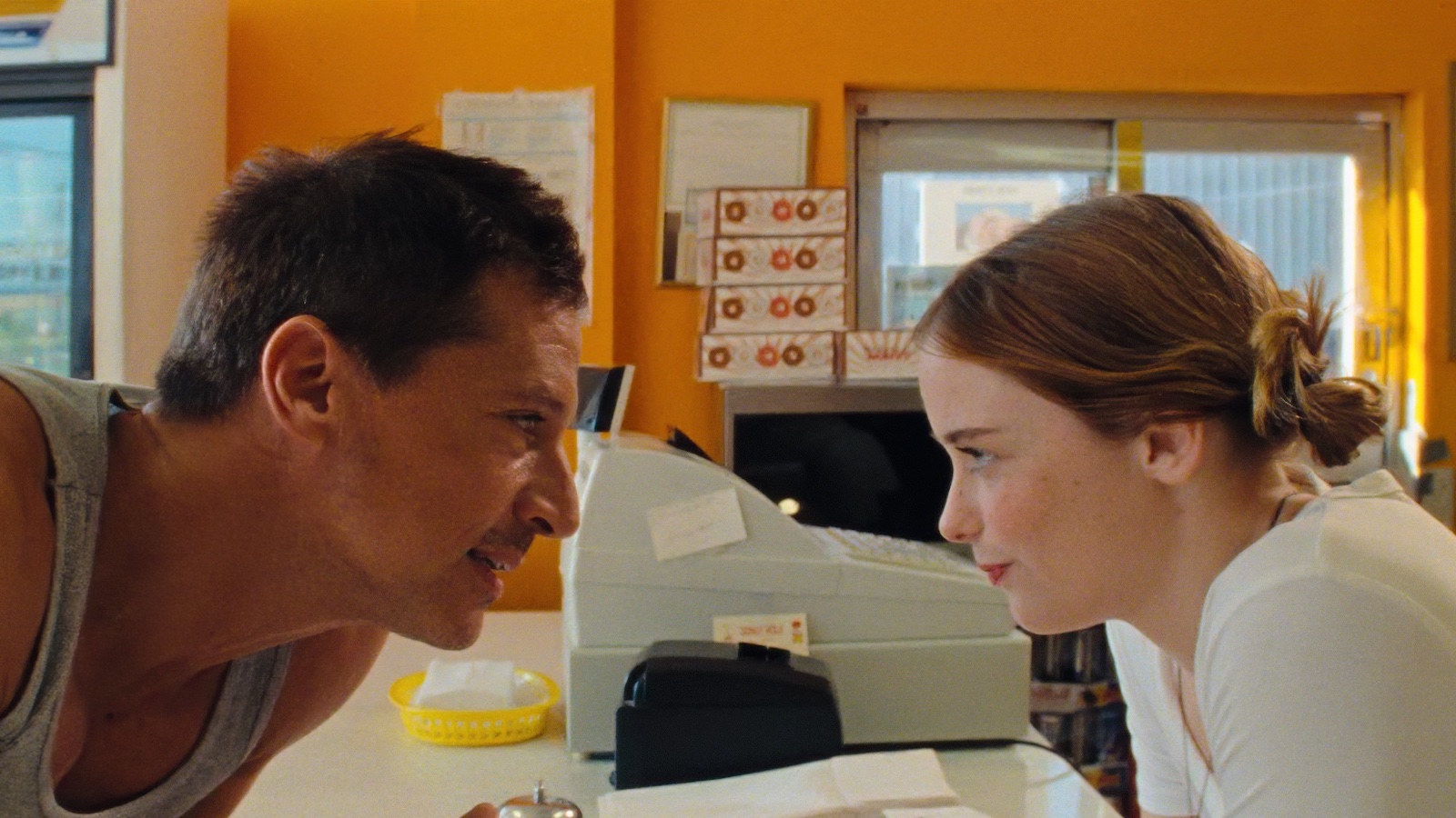 An older man and a younger woman look at each other across the counter at a donut shop, their faces in close profile.