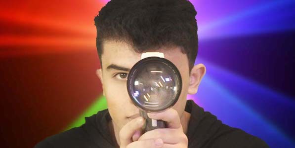 A teenager in dark hair holds a magnifying glass up to his eye as he looks in the camera, a kaleidoscope of colors behind him.