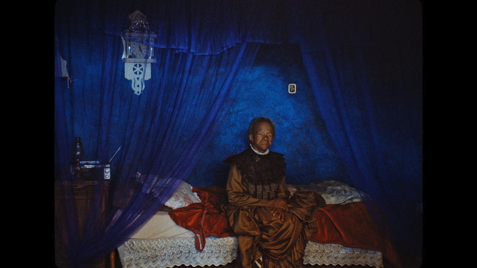 A mournful, elderly woman sits on her bed in a room with deep blue walls.