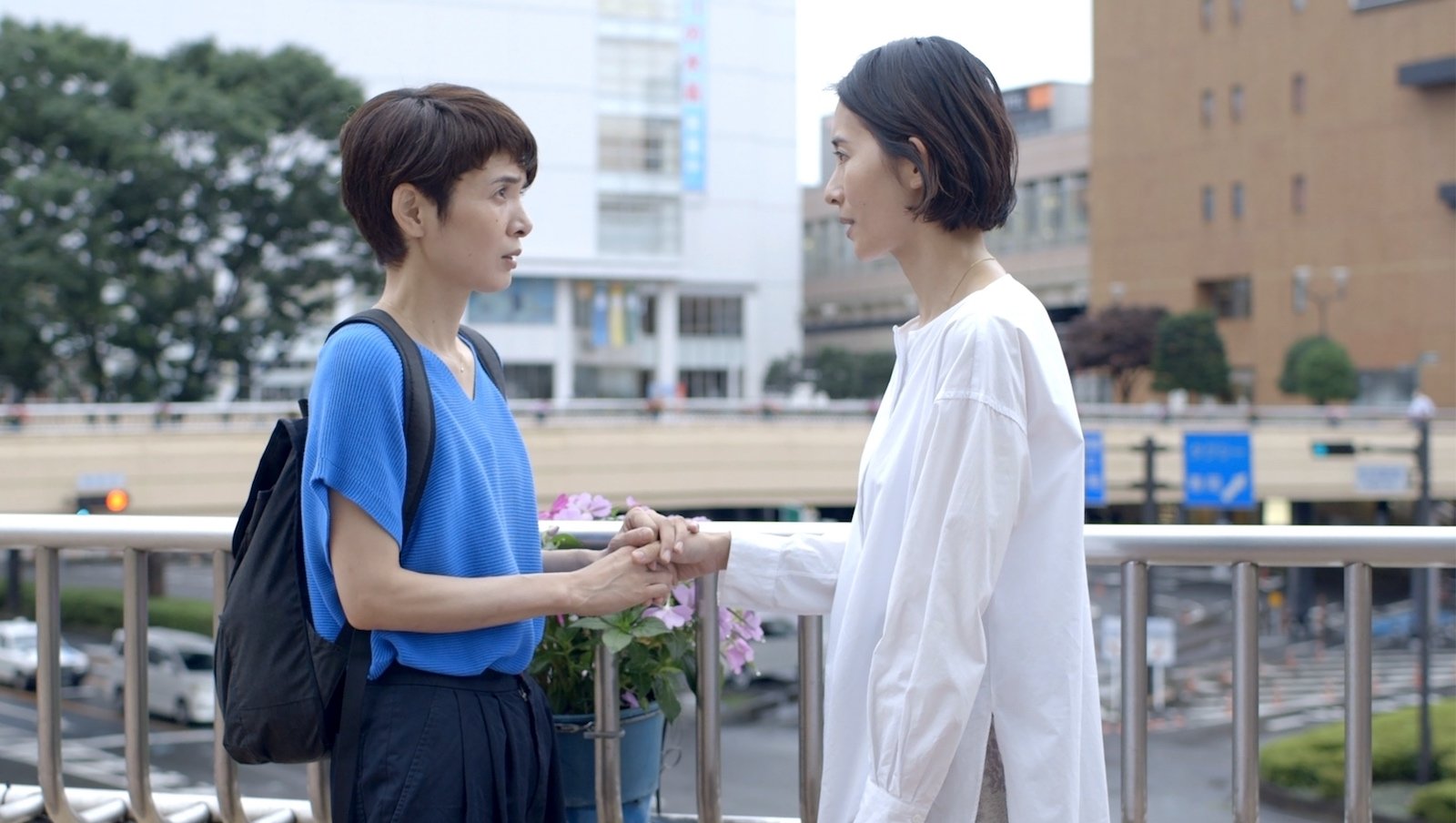 Two women, one in white and one in blue, talk to each other against an outdoor cityscape