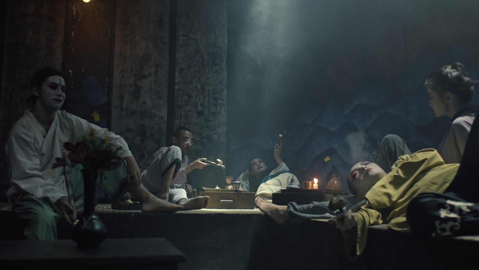 A troupe of actors sprawled around a shadowy den, smoking.