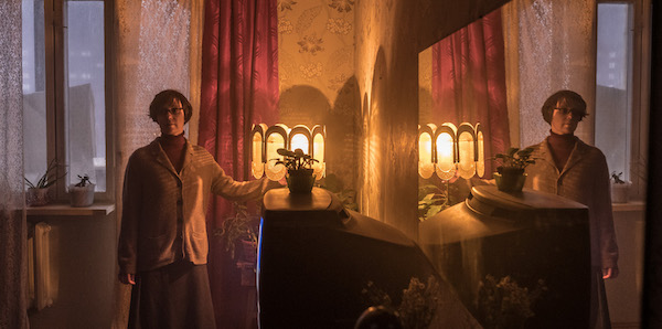A woman stands by a lamp and a plant atop a glowing television screen in a curtained room