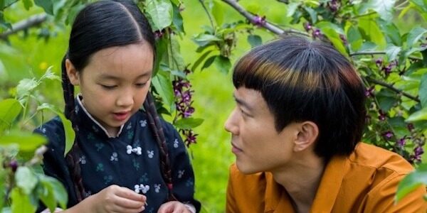 A teenage boy and his little sister crouch in a green forest; she holds a flower as he looks at her.