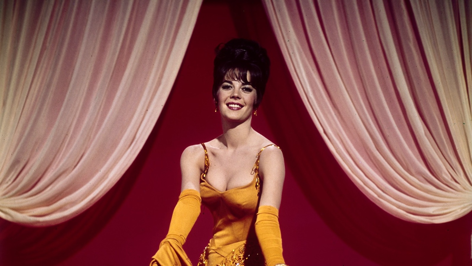 A burlesque performer in a low-cut yellow dress dances, facing the camera.
