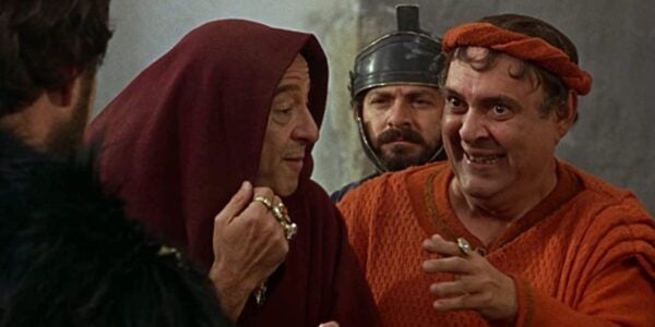 A group of men in ancient Roman garb smile and conspire.