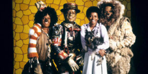 Four cast members of The Wiz, including Dorothy holding her little dog Toto, look at the camera and smile.