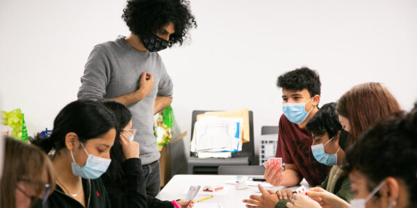 A group of young, masked participants, with one woman standing, surround a table while collaborating on a creative project