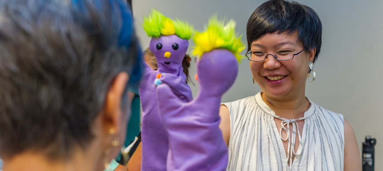 Two women smile and hold purple puppets up to each other