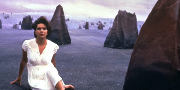 A woman in a white dress sits on a barren landscape surrounded by large, foreboding black rocks