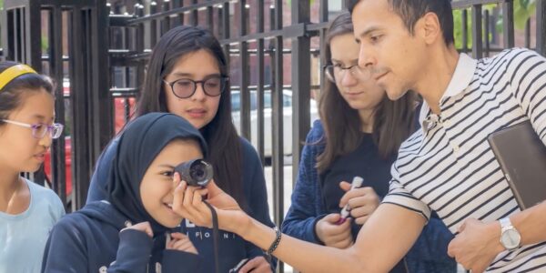 An instructor in a striped shirt holds up a camera lens to the eye of an excited teenage girl, with three teenagers standing behind her
