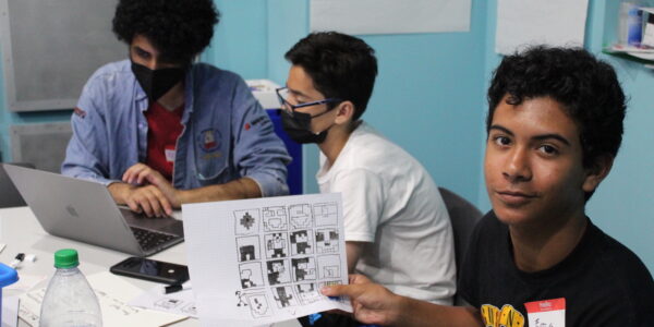 Three teenagers sit at a workshop table; the one in the foreground smiles at camera while he holds up a worksheet with black and white graphic designs on it.