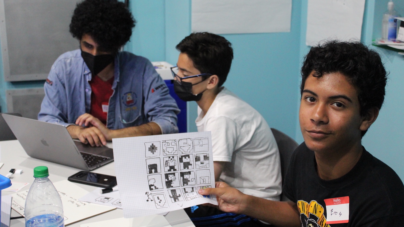 Three teenagers sit at a workshop table; the one in the foreground smiles at camera while he holds up a worksheet with black and white graphic designs on it.