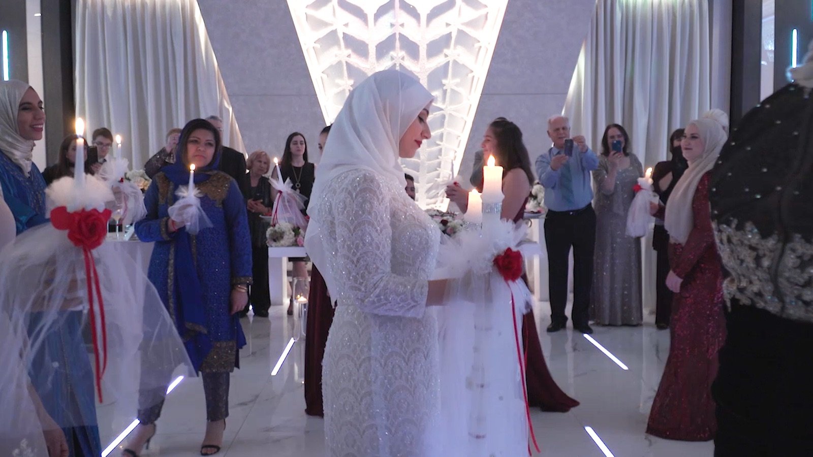 A woman in a white dress and hijab in profile holds a candle in a long hallway with others walking past her