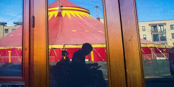 A reflection in a doorway of a man in a wheelchair, set against a red and yellow circus tent