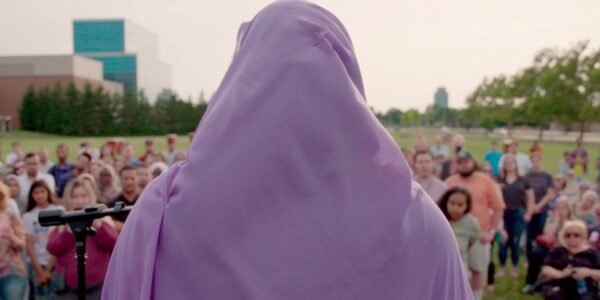 A woman in a purple hijab is seen from behind addressing a crowd in a field