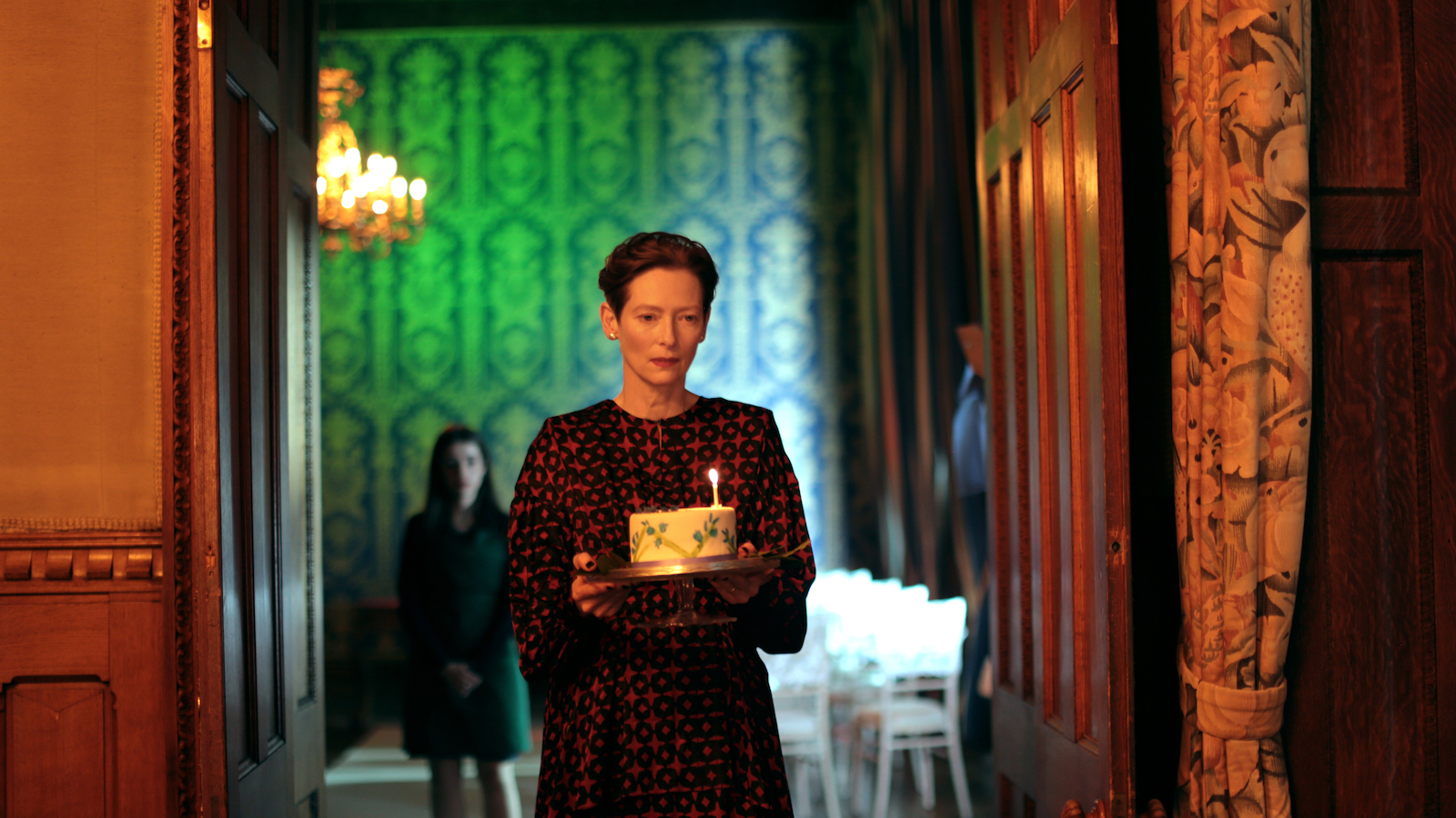 A woman in a hallway holds a birthday cake against a green wallpaper background