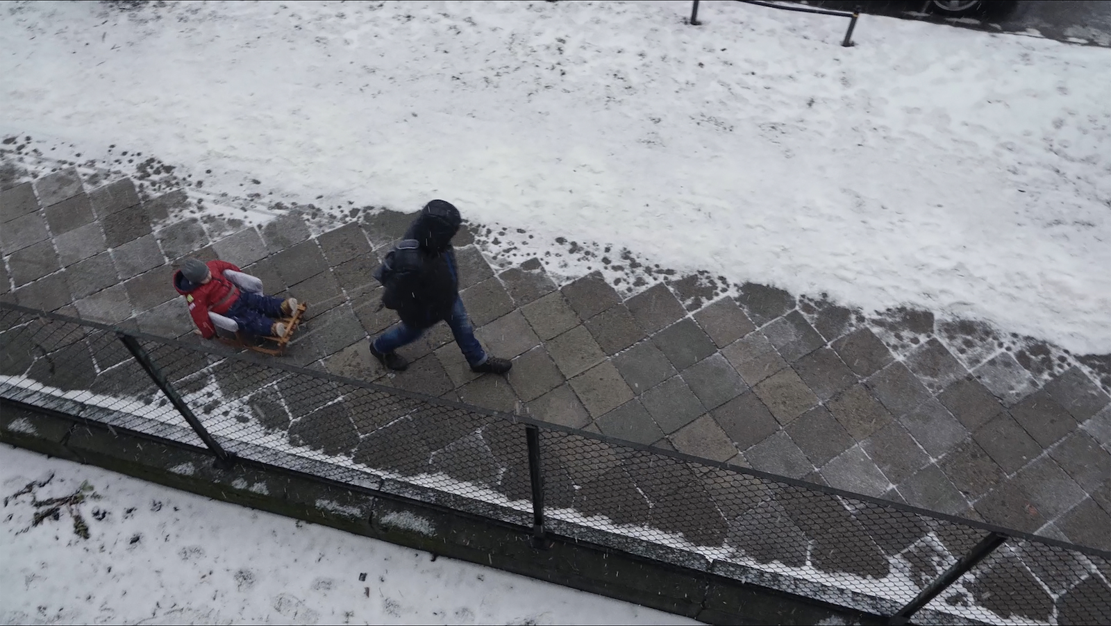 An adult figure pulls a child in a small sled on a wintry street as seen from a high balcony.